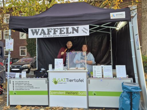 Zwei Personen stehen am Waffelstand mit Menü und Werbung in einem Festival-Setting.