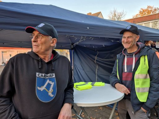 Zwei Männer stehen neben einem Tisch, einer trägt eine Warnweste, der andere ein schwarzes Sweatshirt.