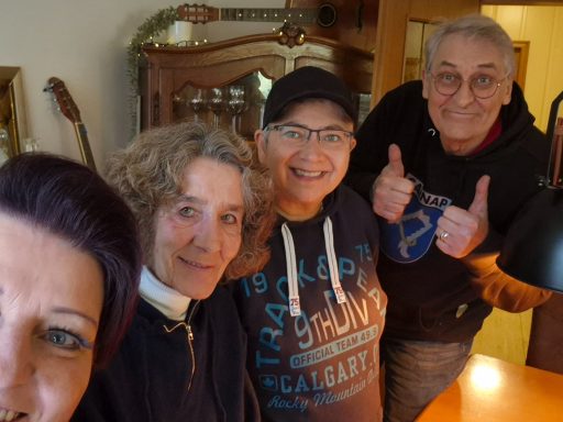 Vier Personen lächeln in einem gemütlichen Raum, zwei zeigen Daumen hoch.