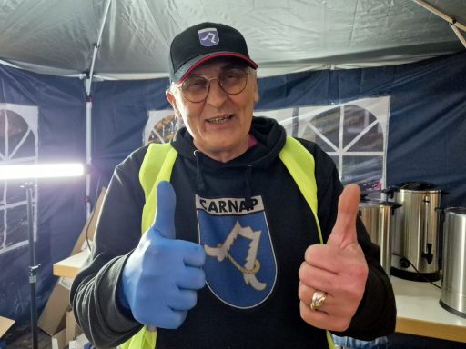 Lächelnder Mann mit blauen Handschuhen, Daumen hoch, in einer Zeltumgebung.