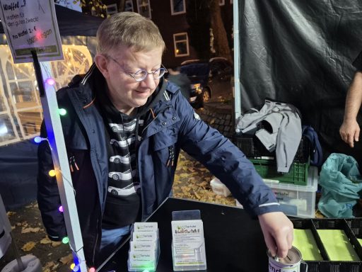 Mann mit Brille und blauer Jacke reicht eine Packung über einen Tisch auf einem Markt.
