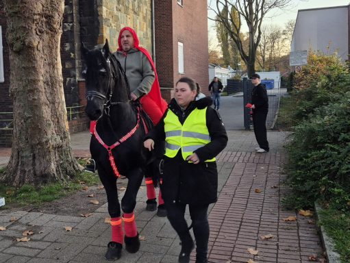 Eine Person in rotem Umhang reitet ein Pferd, während eine Frau in Warnjacke daneben geht.
