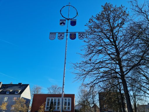 Maibaum mit Wappen und Fahnen vor klarem blauen Himmel und Bäumen.
