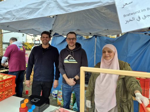 Drei Personen stehen an einem Stand auf einem Markt, umgeben von bunten Kisten.