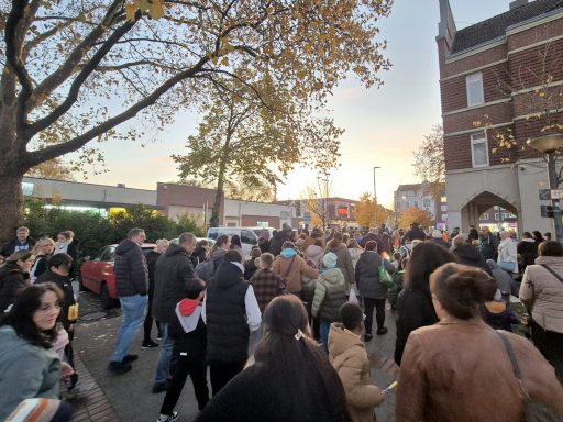 Menschenmenge auf einer Straße, umgeben von Bäumen und Gebäuden, bei Sonnenuntergang.