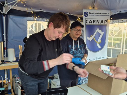 Zwei Personen an einem Stand, eine hält ein Gerät, die andere bereitet etwas vor.
