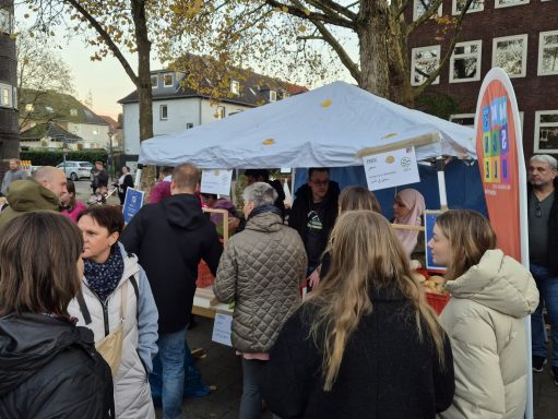Menschen um einen Stand auf einem Markt, im Hintergrund ein Zelt und Bäume.