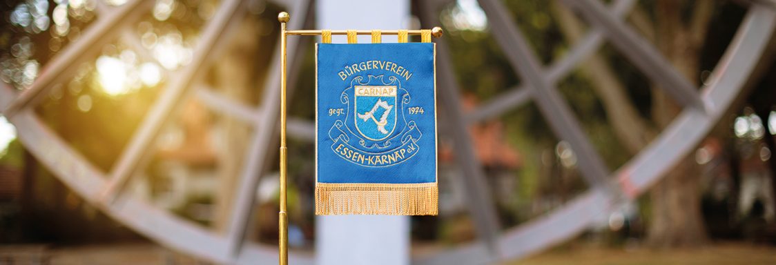 Blaues Wappen-Fahne vor verschwommenem Hintergrund eines Metallmonuments.