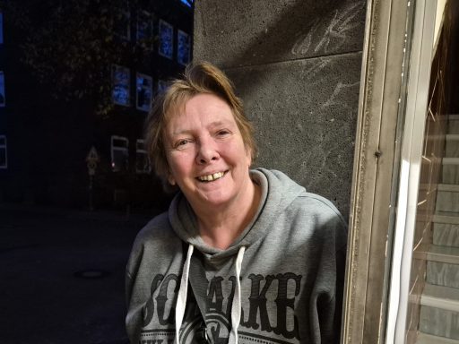 Frau mit kurzer blonder Frisur und grauem Sweatshirt, lächelt freundlich.