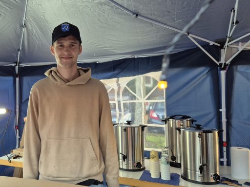 Junger Mann in einem Zelt, lächelnd, neben Kaffeekannen und einem Tisch.