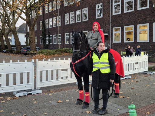 Ein Reiter in rotem Gewand auf schwarzem Pferd, begleitet von einer Person in Warnweste.